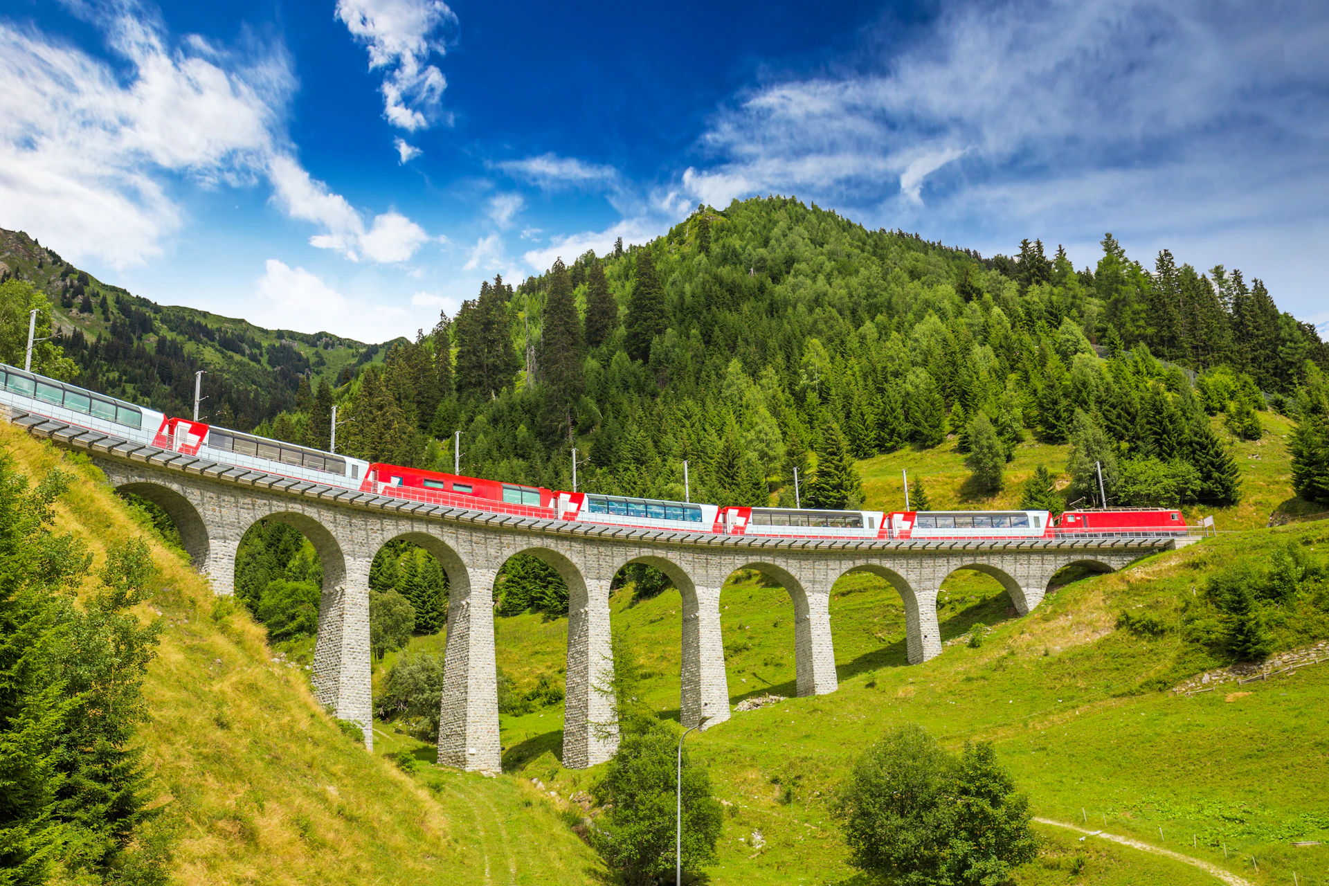 A red passenger train crosses a tall, arched stone viaduct amidst lush green hills under a bright blue sky.