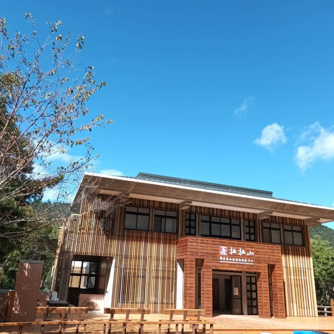 A two-story building with wooden accents under a blue sky, surrounded by trees and featuring a clear entrance.