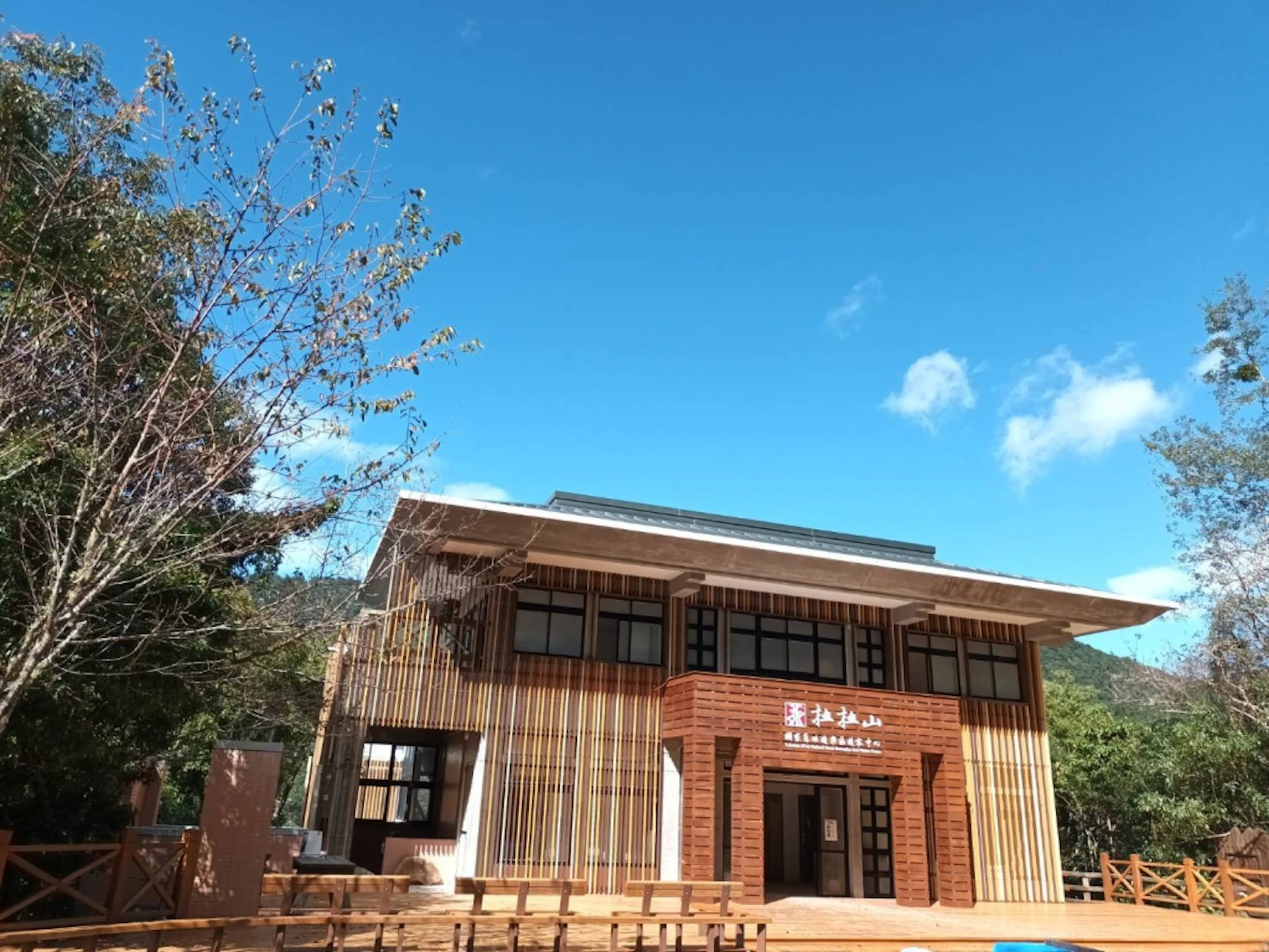 A modern two-story wooden building with large windows and decorative vertical slats, set under a blue sky with trees nearby.