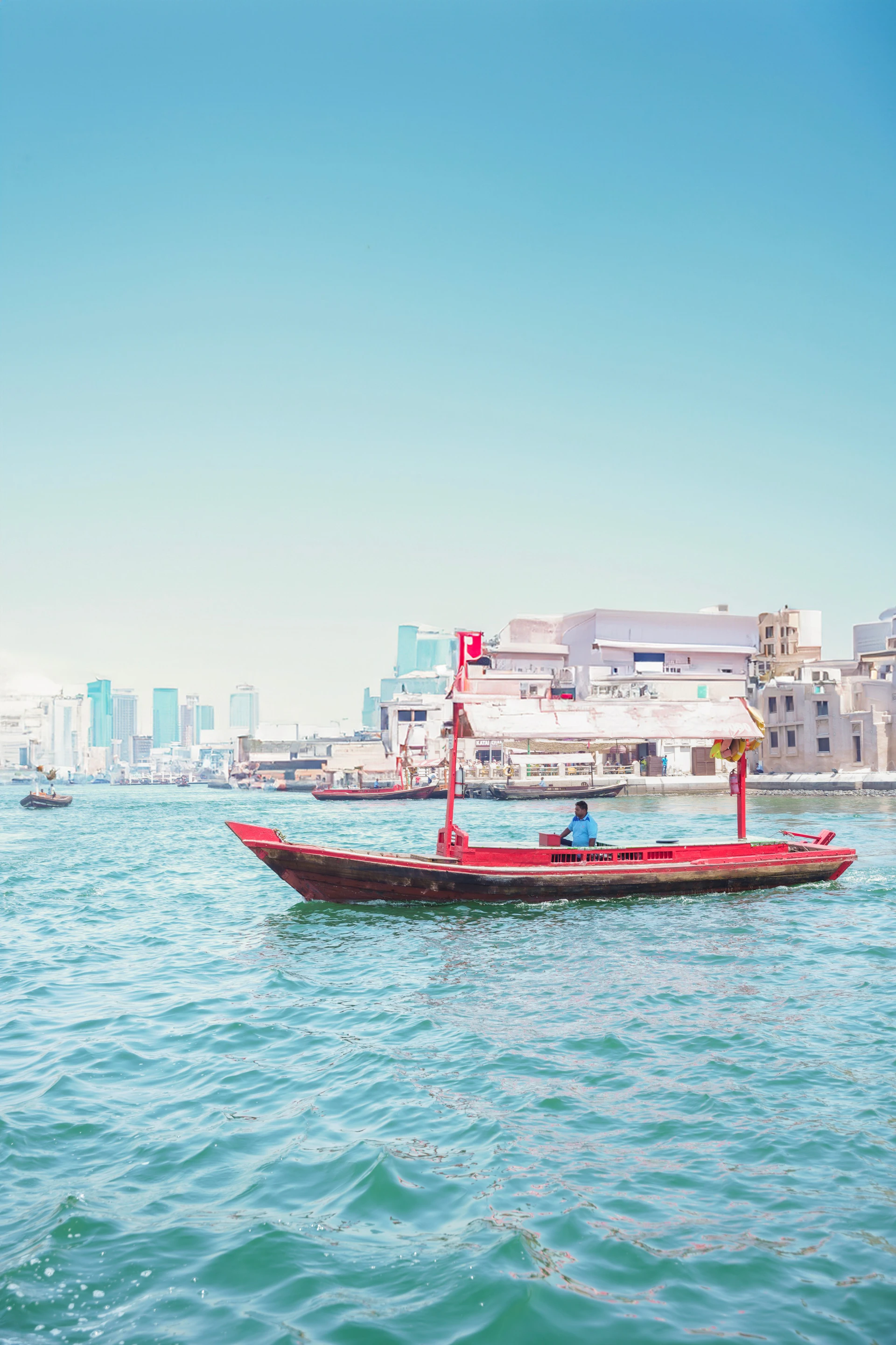 A person steering a traditional red boat on a bright, sunny day with a city skyline and waterfront buildings in the background.