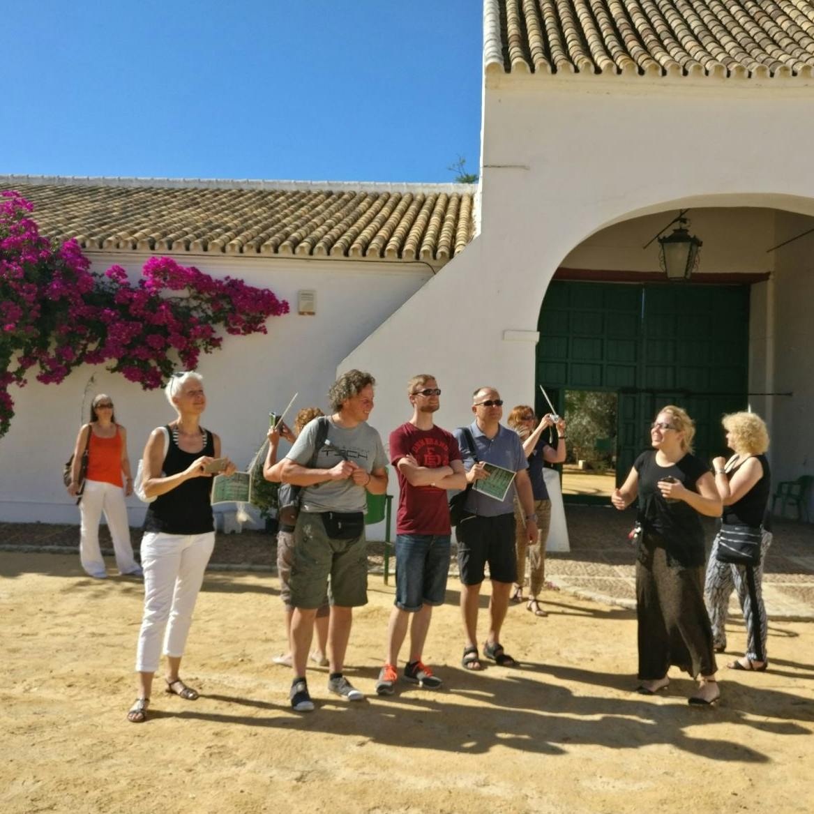 Skupina lidí stojí před bílou budovou s klenutými vchody, pod jasnou modrou oblohou, poblíž pulzující bougainvillea.
