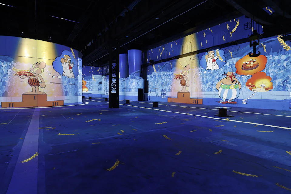 A colourful projection display of cartoon characters on the walls and columns in a dark exhibition space with a blue floor.
