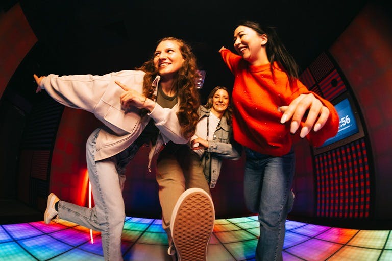 Tre unga kvinnor dansar glatt på ett pulserande, färgstarkt upplyst golv i ett mörkt rum med neondialekter.