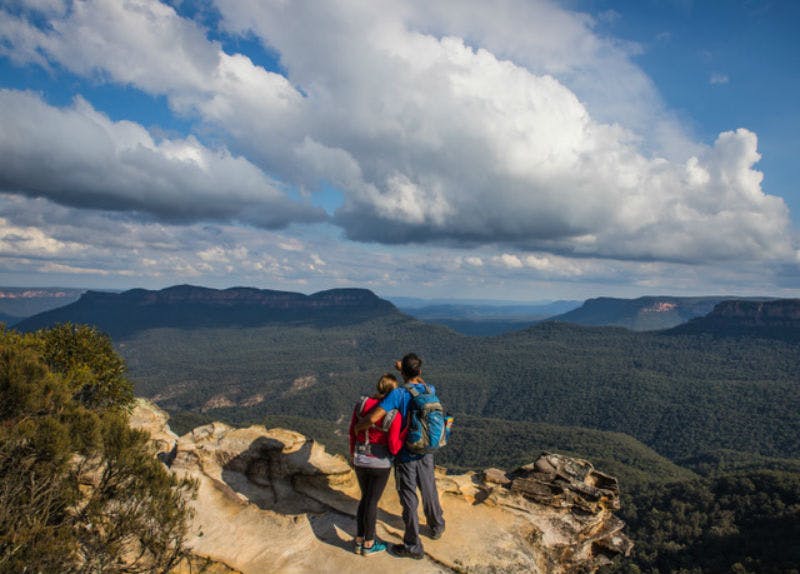 Two people with backpacks stand on a rock ledge overlooking a vast, forested mountain range under a cloudy sky.