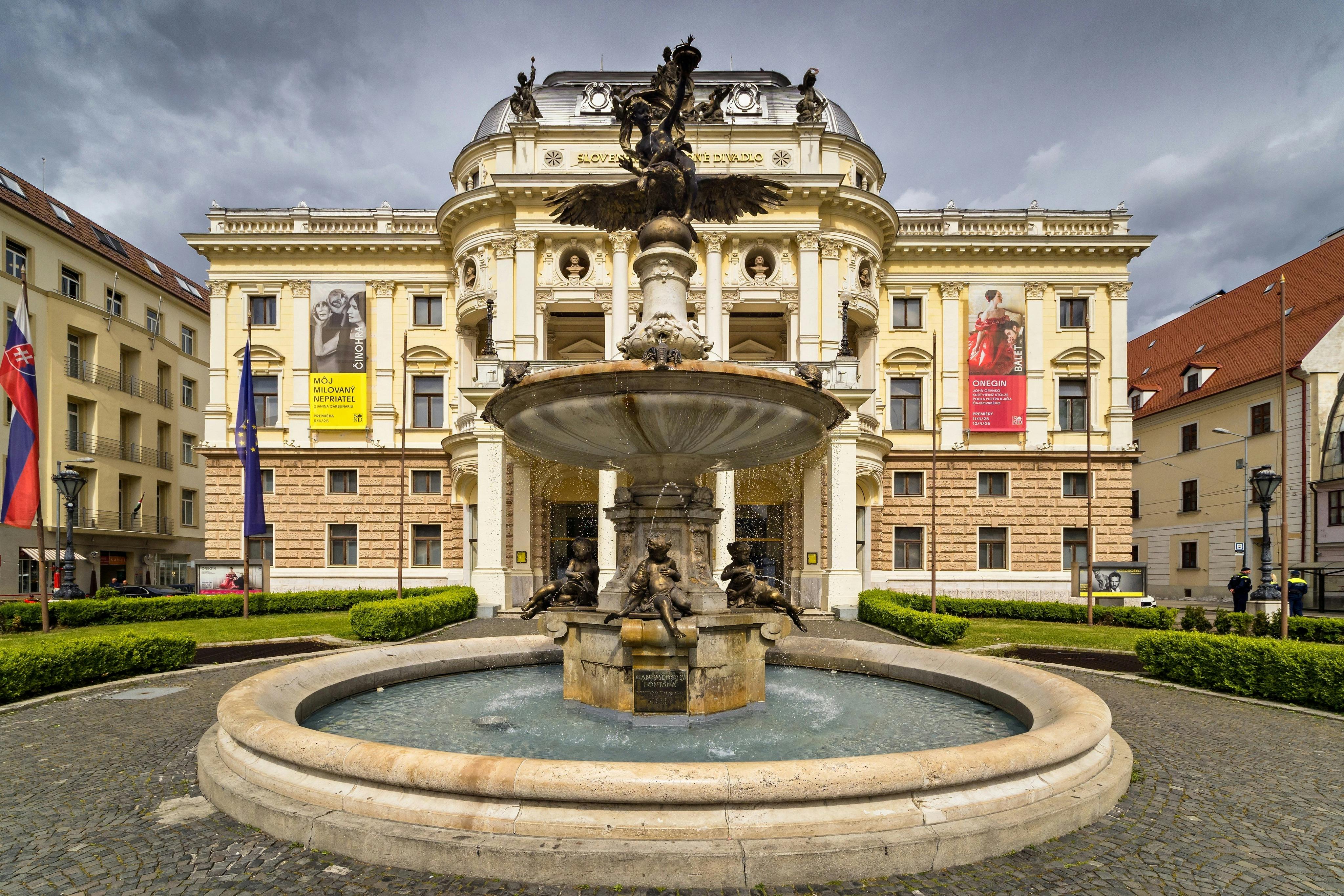 A decorative fountain in front of an ornate building with columns and banners under a cloudy sky.