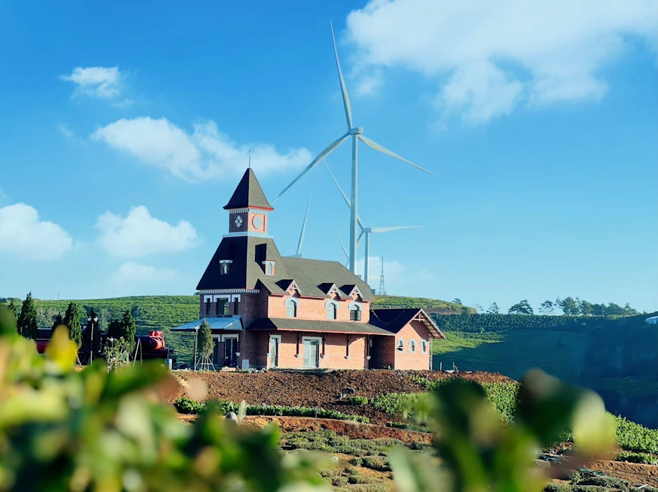 A brick house with a clock tower surrounded by greenery and wind turbines in the background under a blue sky.