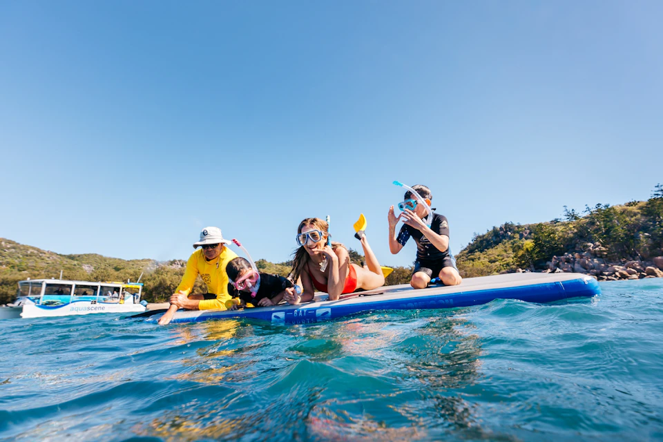 Four people in snorkeling gear on a paddleboard in clear blue water under a sunny sky, near a hilly, forested shoreline.