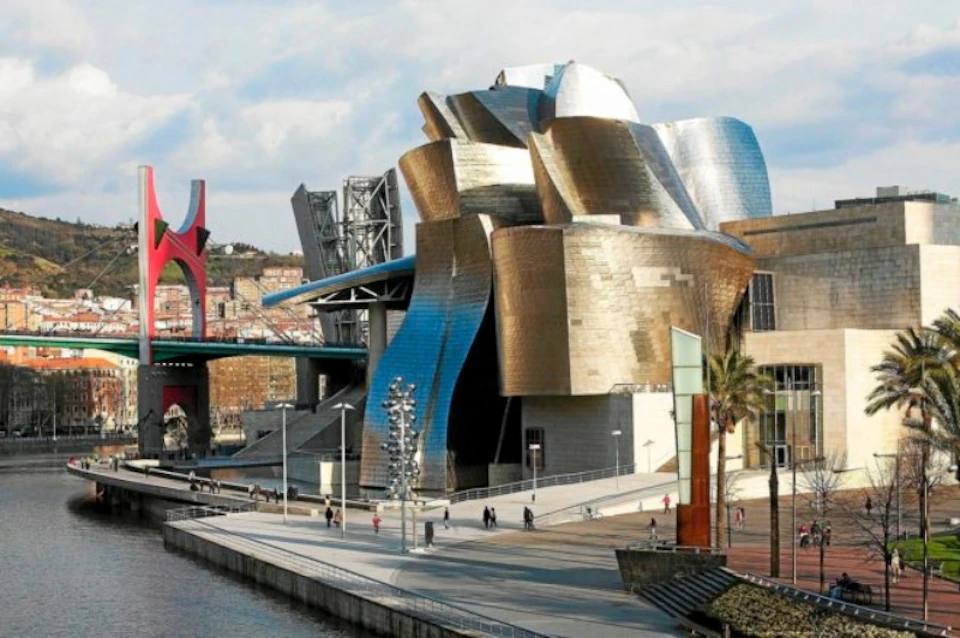 Modern metallic building with curved, reflective surfaces beside a riverfront walkway. People walk on the path below.
