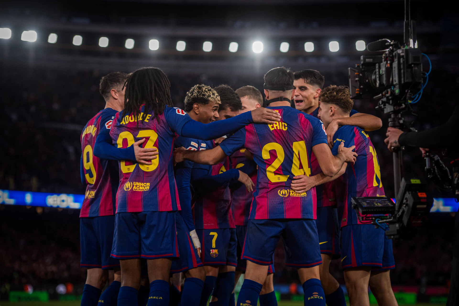 A group of soccer players in blue and red uniforms celebrating on a field under bright stadium lights.