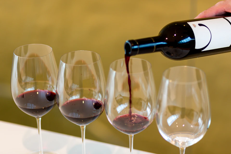 Red wine being poured from a bottle into one of four wine glasses, with some glasses partially filled.