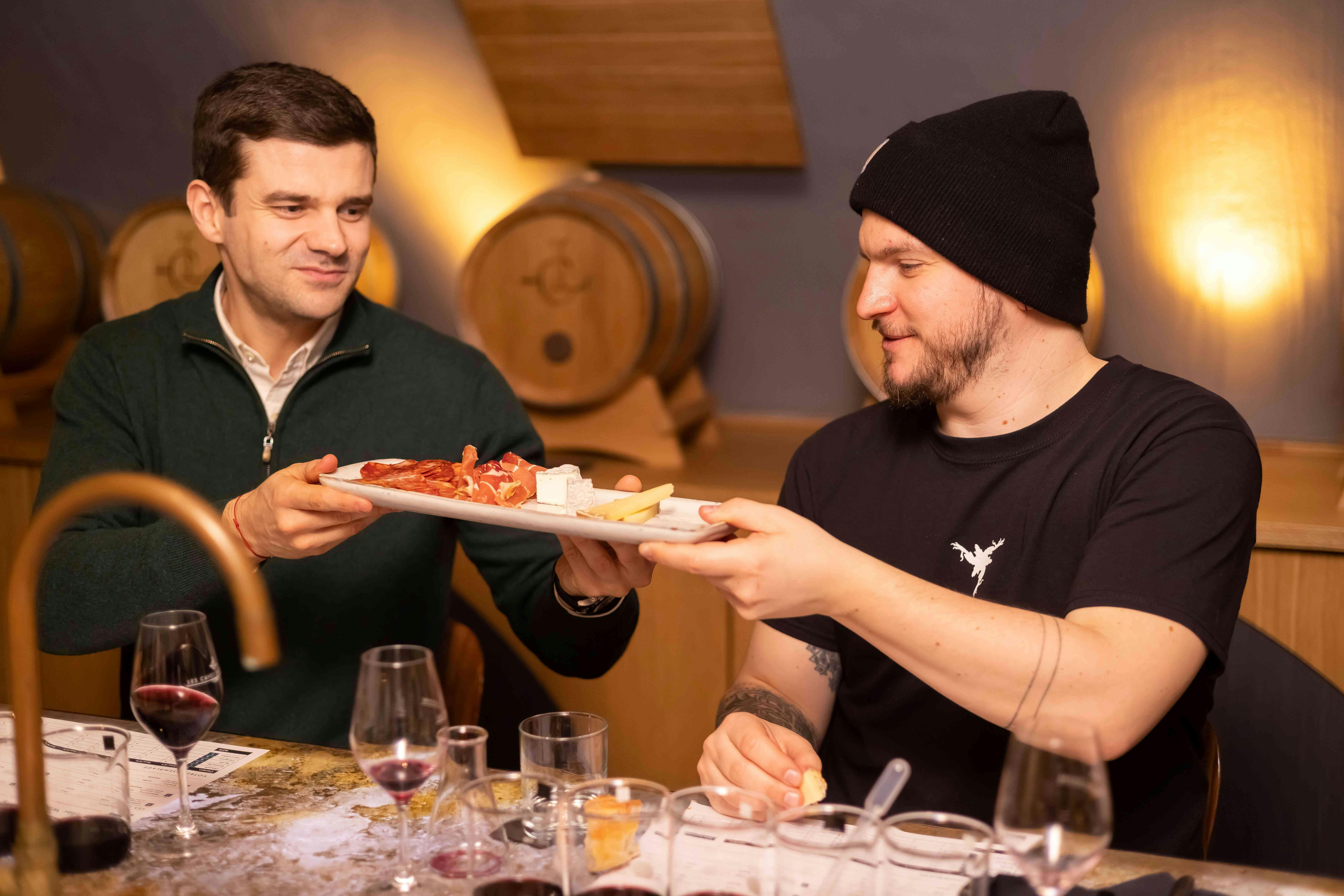 テーブルに座った2人の男性が、ワイングラスと樽を背景にしたシャルキュトリーの盛り合わせのプレートを通り過ぎています。
