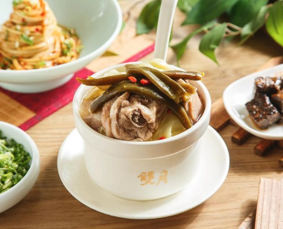 A bowl of soup with meat, green beans, and garnishes, served in a white container on a wooden table with other dishes nearby.
