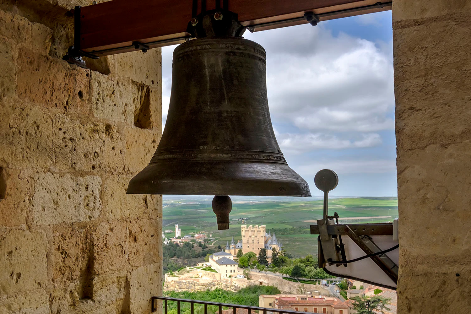 Large bronze bell in a stone tower with a view of a distant castle and countryside under a partly cloudy sky.
