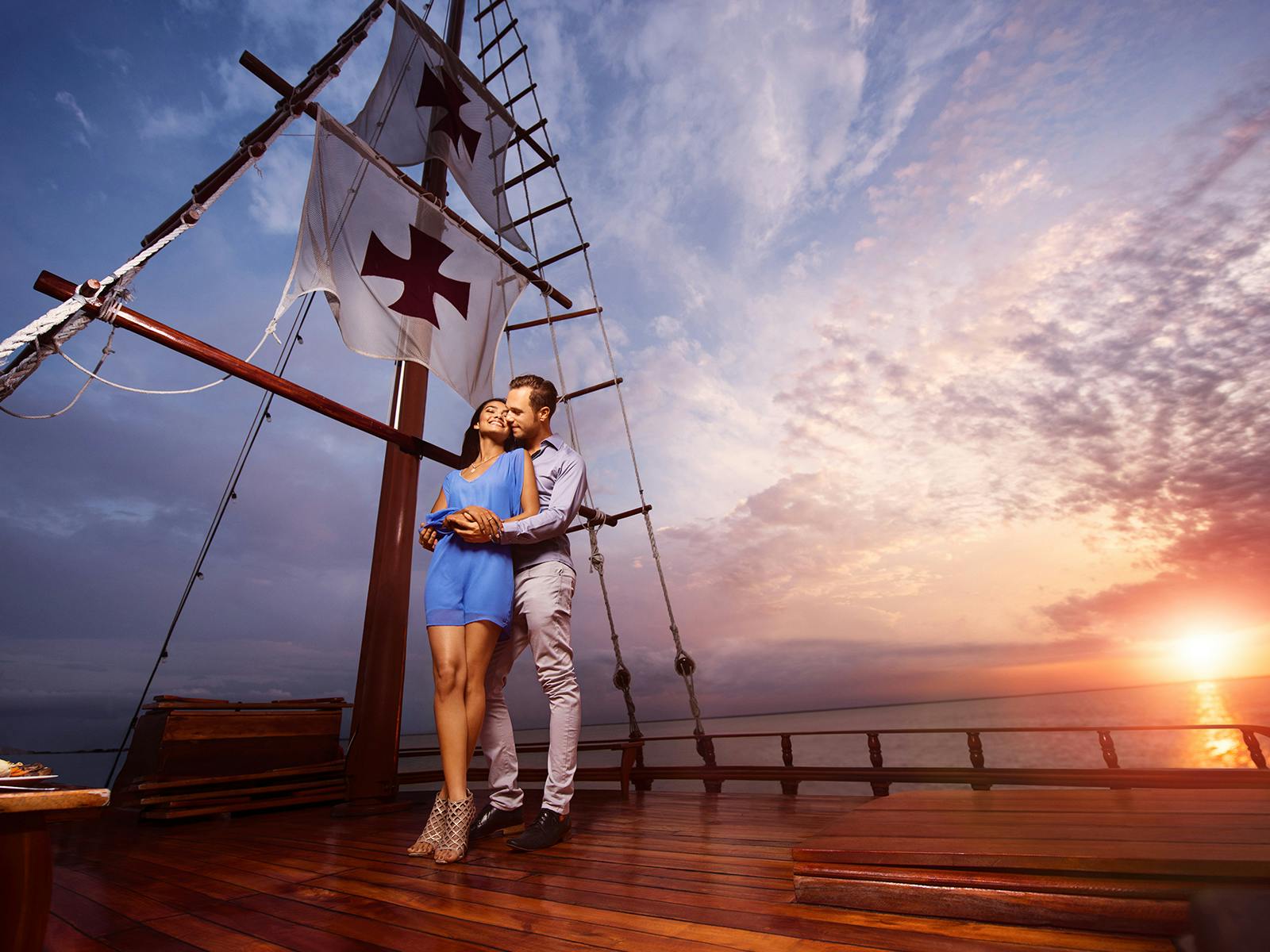 Una coppia si abbraccia sul ponte di una nave di legno con grandi vele e un tramonto sullo sfondo.