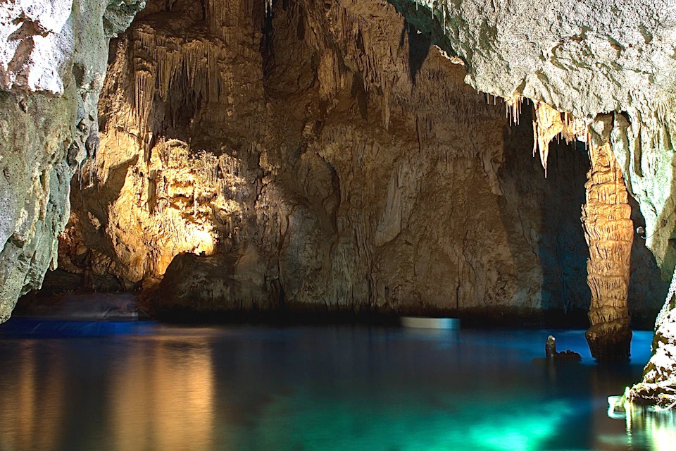 An illuminated underground cave with stalactites and clear water reflecting blue and green light.