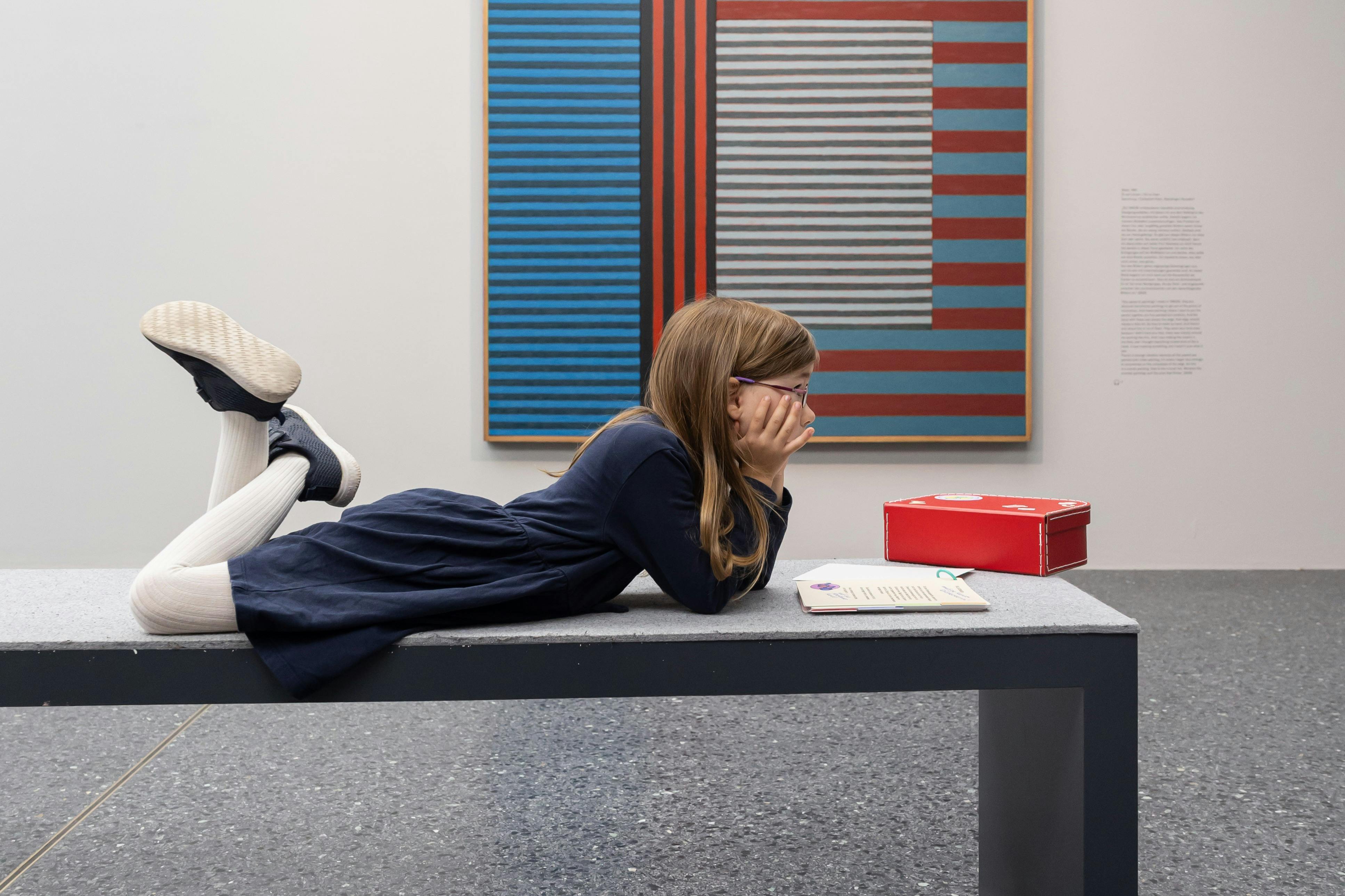 한 어린이가 갤러리의 벤치에 누워 빨간색 상자 근처의 수첩을 보고 있습니다. 벽에 줄무늬 추상화가 걸려 있습니다.