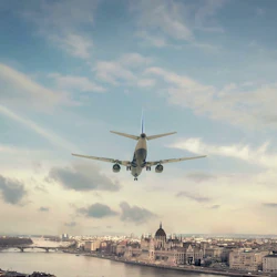 Flygplan som flyger över en stad med en flod, broar och historiska byggnader under en delvis molnig himmel.