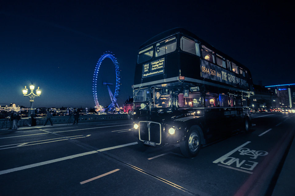 A black double-decker bus on a city street at night with the London Eye illuminated in blue in the background.