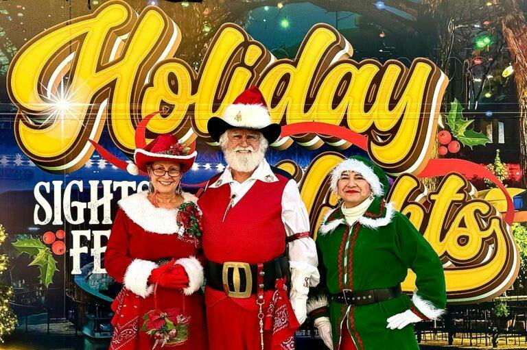 Three people in festive holiday costumes posing in front of a "Holiday" sign with decorative lights.