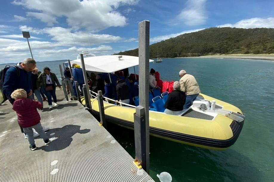 A group of people boarding a yellow inflatable boat docked at a pier, with a forested shoreline in the background.
