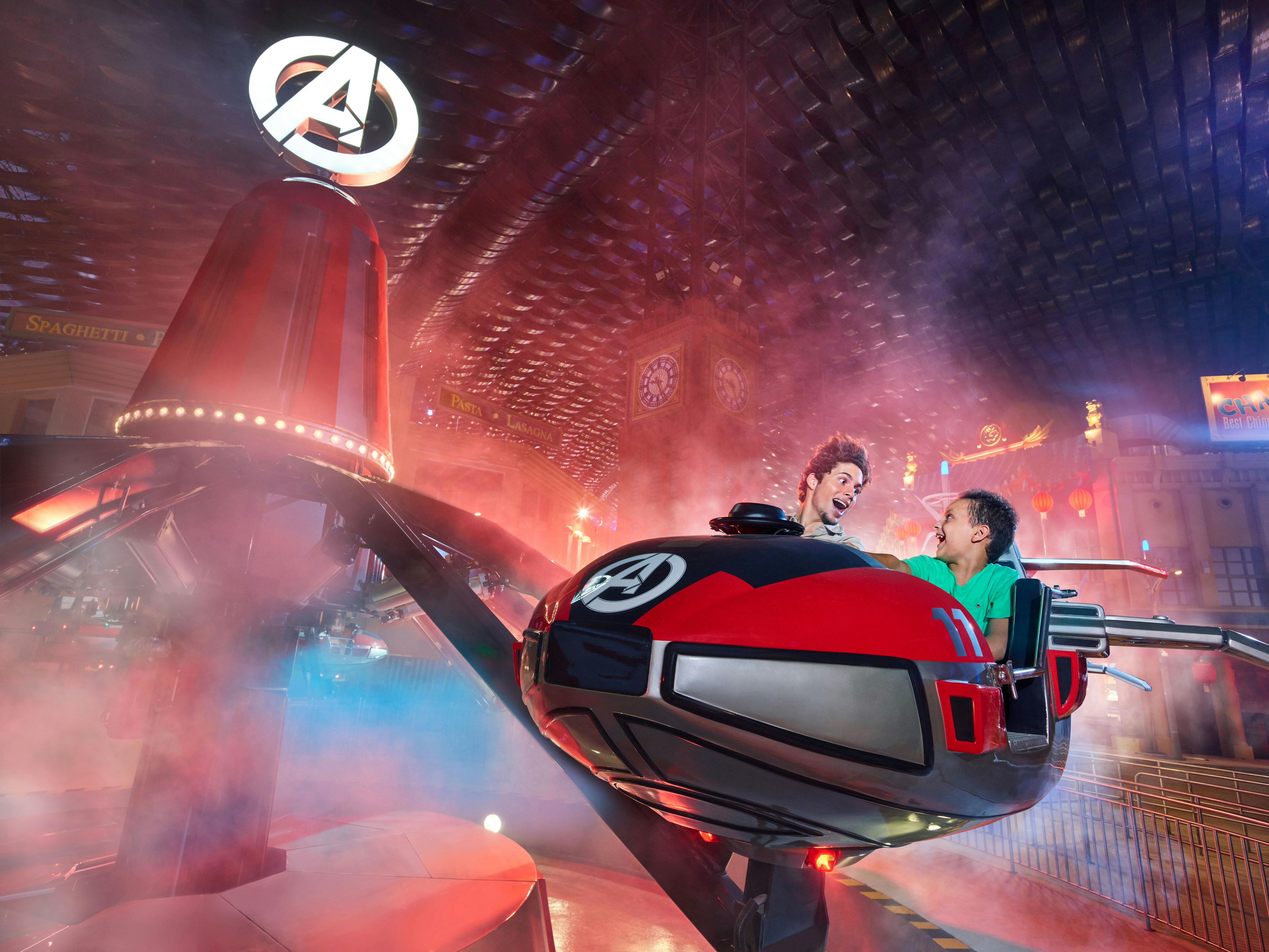 Δύο άνθρωποι οδηγούν με ενθουσιασμό μια περιστρεφόμενη βόλτα διασκέδασης με θέμα τους Εκδικητές σε ένα εσωτερικό, φωτισμένο περιβάλλον.