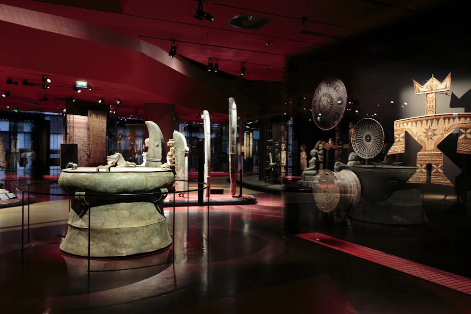 A dimly lit museum exhibit showcasing ancient sculptures and artifacts, with reflective glass and spotlighted displays.