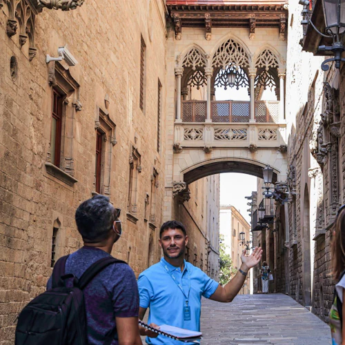 En turguide gestikulerar mot en gotisk bro som förbinder två byggnader i en smal, historisk gata, med två turister som tittar på.