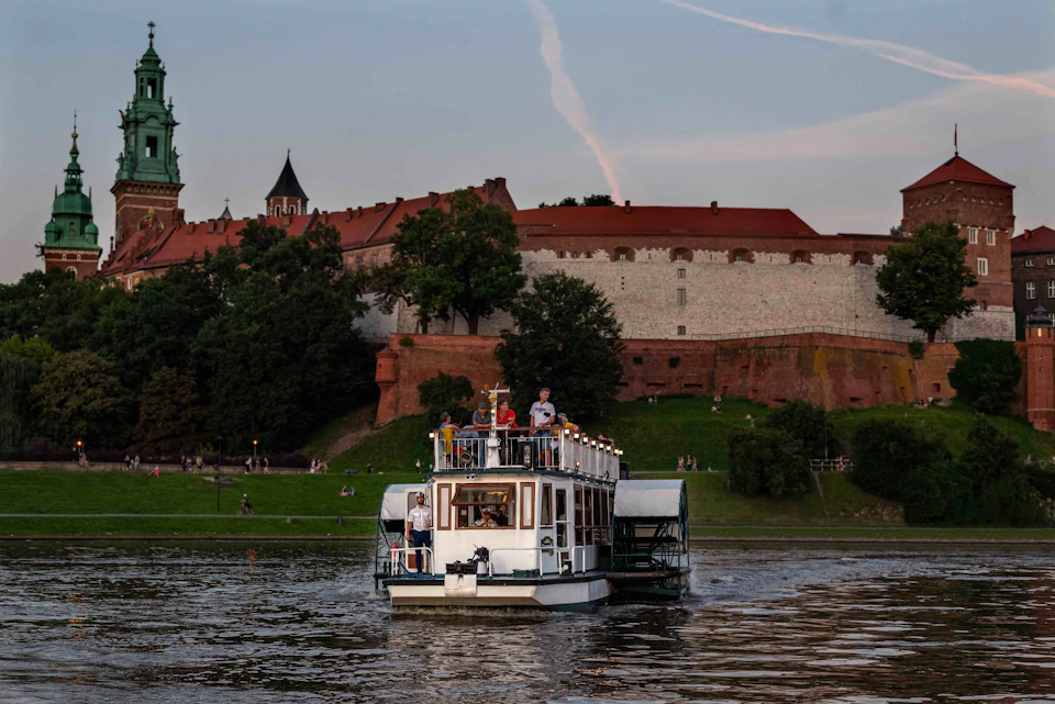 A small tour boat with passengers cruises along a river with a historic castle and lush greenery in the background at dusk.