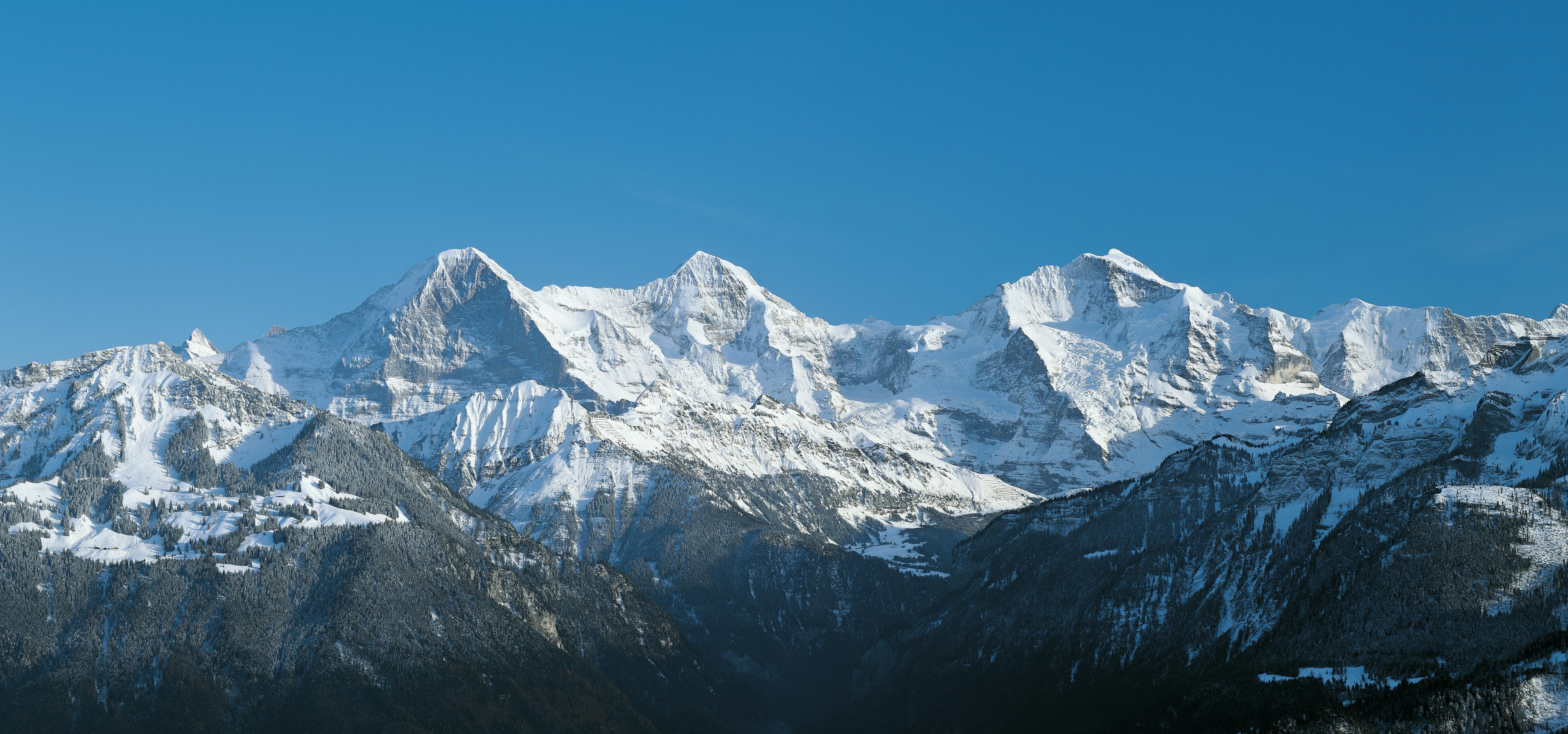 Χιονισμένα βουνά κάτω από έναν καταγάλανο ουρανό, με χαμηλότερες δασωμένες πλαγιές που καλύπτονται εν μέρει από χιόνι.