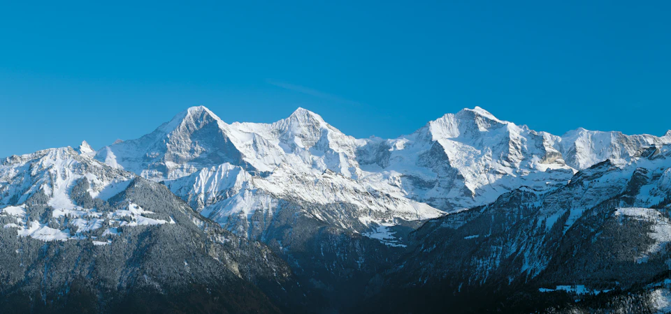 Snow-covered mountains under a clear blue sky, with lower forested slopes partly covered in snow.