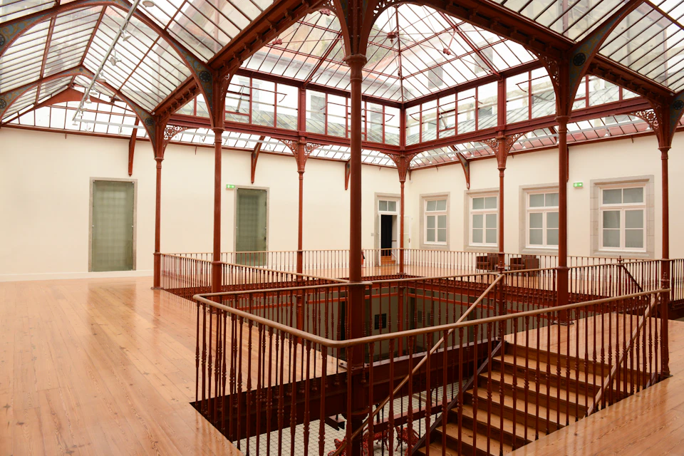 A spacious indoor atrium with wooden floors, red iron railings, and a glass ceiling, surrounded by doorways and windows.