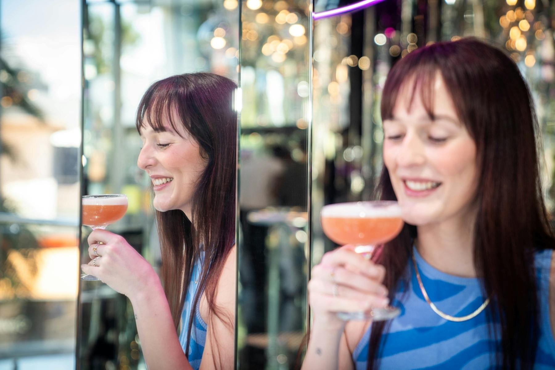 青いストライプの服を着た長い髪の女性がカクテルを飲みながら微笑んでいます。鏡のような壁に映っています。