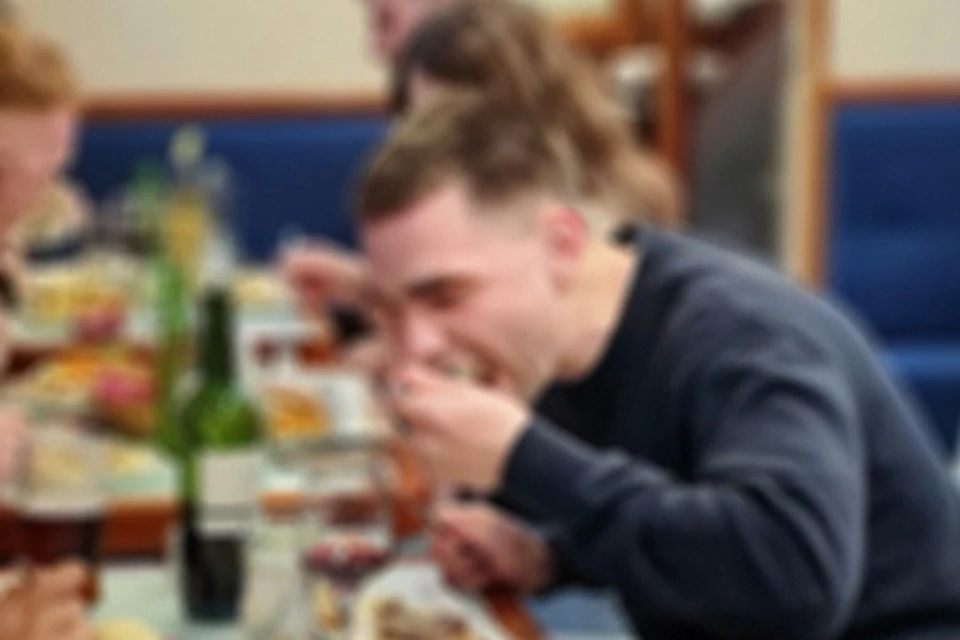 A woman smiling at the camera while dining with others at a restaurant. Plates of food and glasses of wine are on the table.