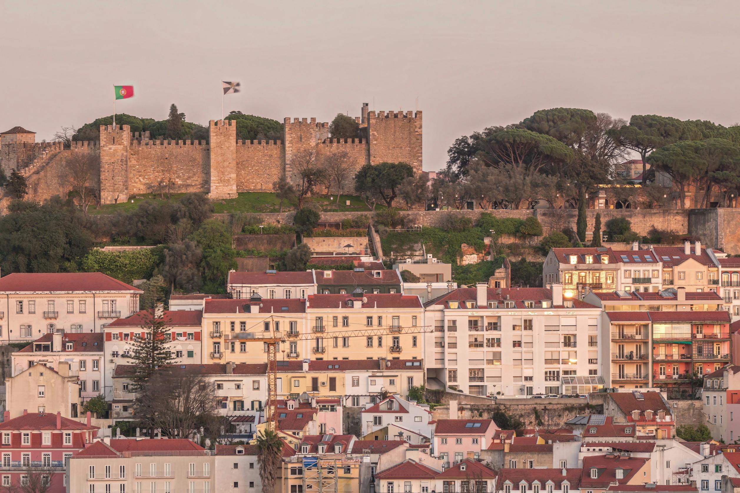 A historic castle with battlements overlooks a hillside neighborhood with red-roofed houses and trees, during sunset.
