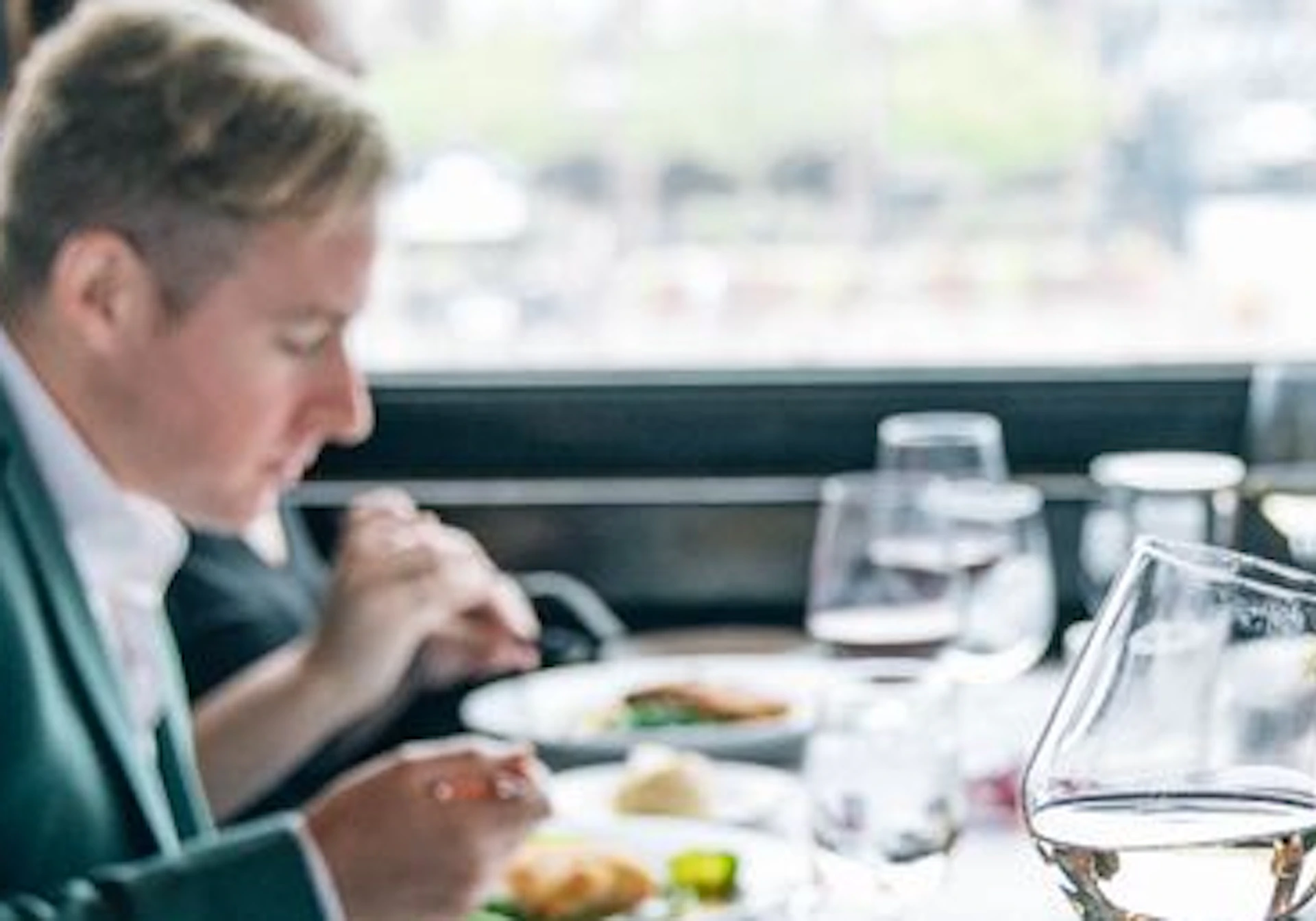 People sitting at a dining table by a window, toasting with wine glasses, with plates of food and cutlery in front of them.