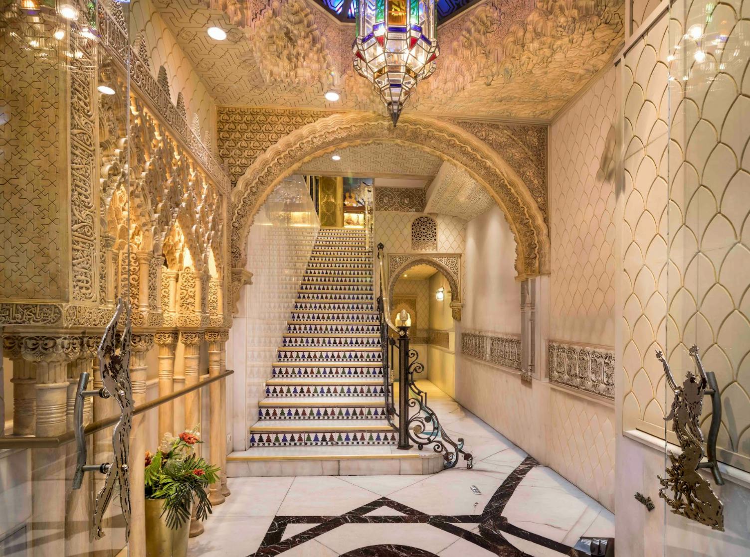 Ozdobna klatka schodowa z misternymi łukami, wzorzystymi płytkami i kolorowym żyrandolem w bogato zdobionym wnętrzu.