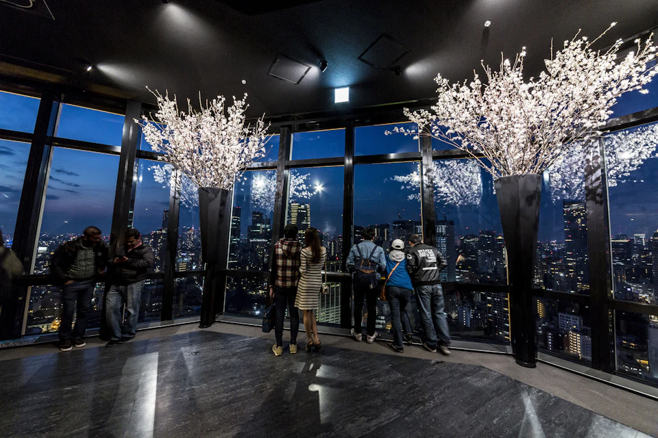 People standing by large windows, looking at a city skyline at night. There are large vases with white flowers on either side.