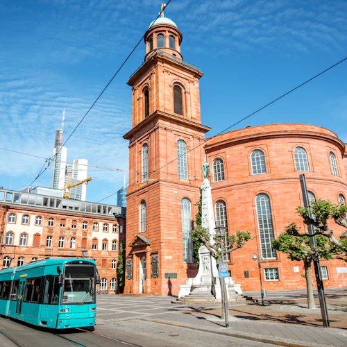 En turkos spårvagn passerar en historisk kyrka i rött tegel och ett monument under en klarblå himmel i en urban miljö.