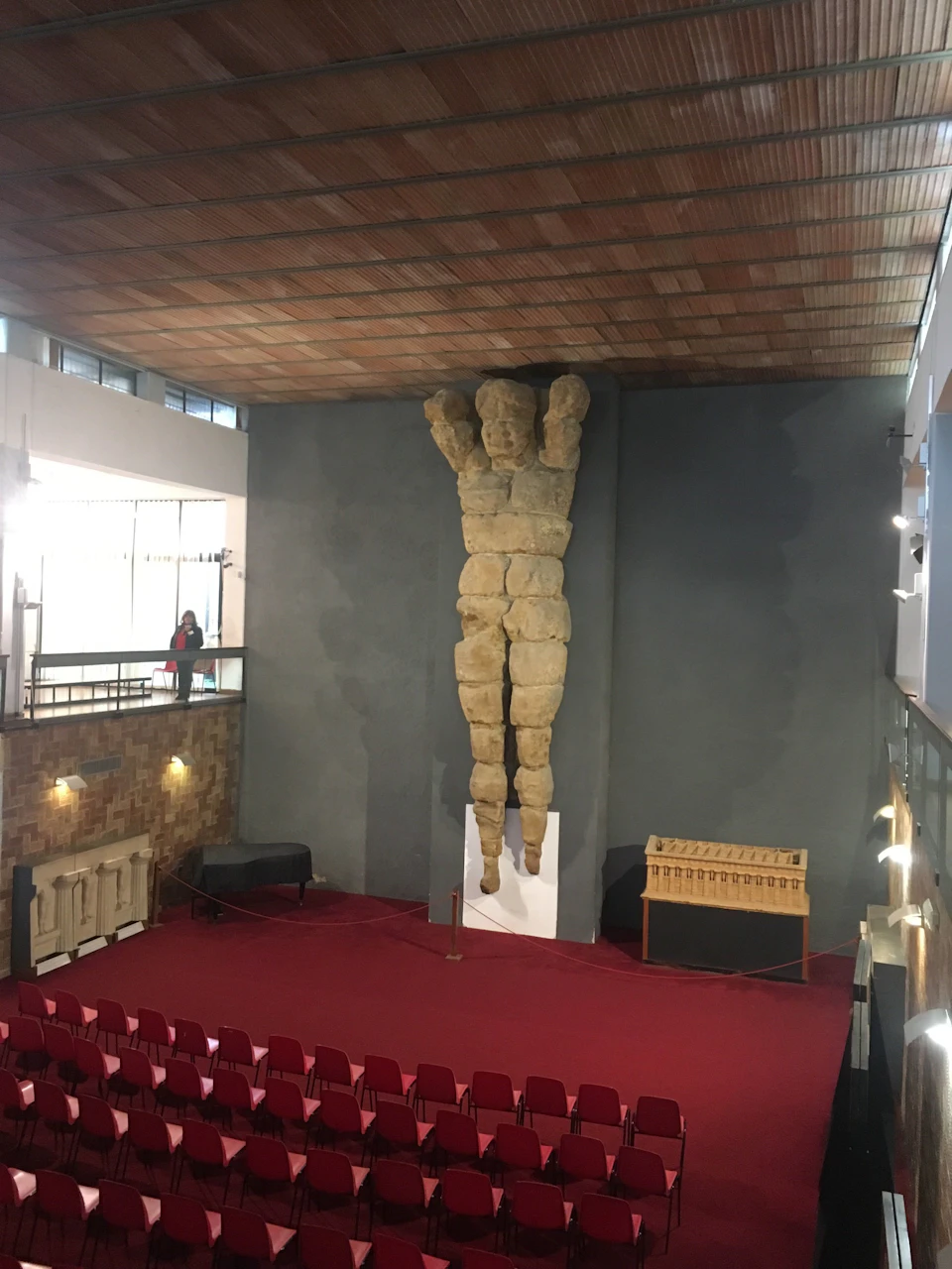 Large stone statue embedded in a wall, arms raised, overlooking rows of red chairs in a spacious room with a wooden ceiling.