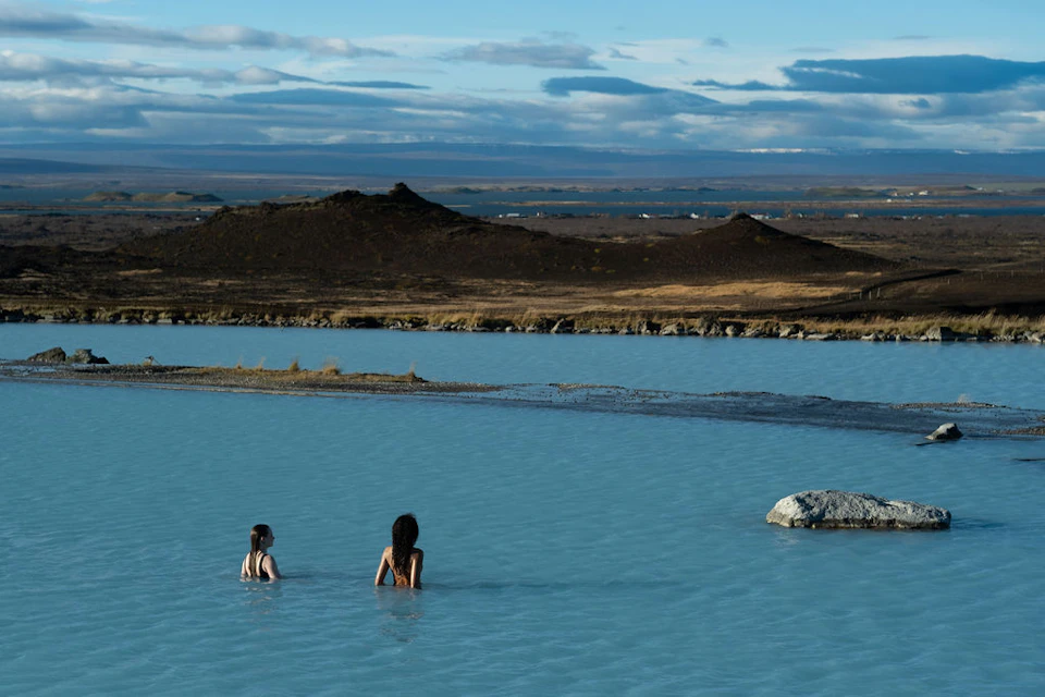 Two people in swimsuits wade in a blue geothermal pool surrounded by a rocky landscape under a cloudy sky.