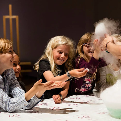 Barn ler och sträcker sig entusiastiskt mot skummande bubblor som skapas av en person i en röd skjorta vid ett bord.