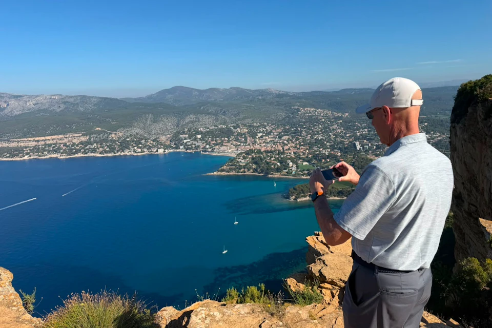 A man wearing a cap and sunglasses takes a photo with his phone from a cliff overlooking a bay and a coastal town.