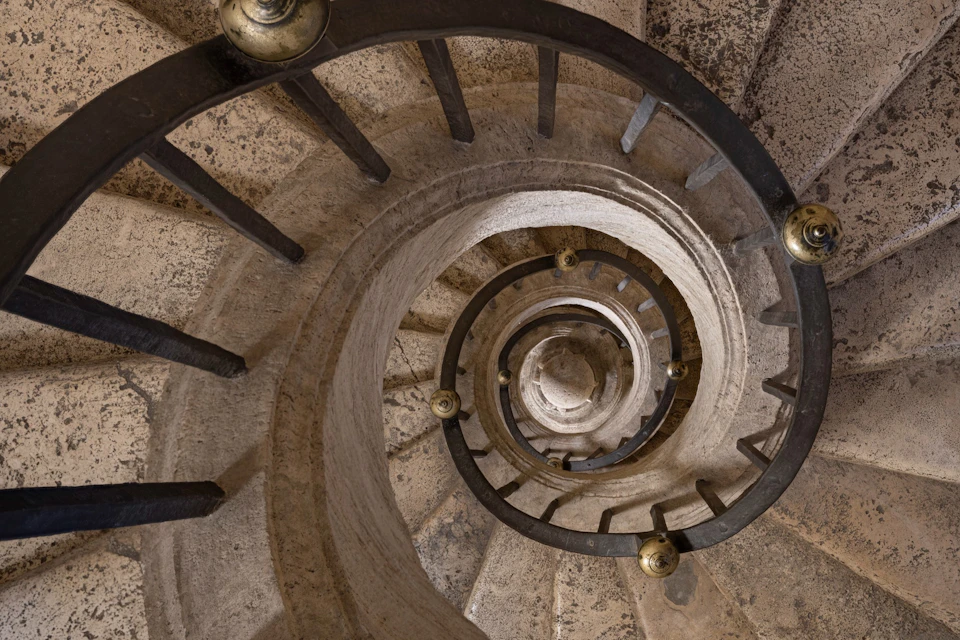 Spiral stone staircase with black metal railings, viewed from above, showing multiple descending levels.