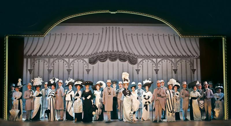 A group of people dressed in formal, period-style clothing on stage with a decorative backdrop.