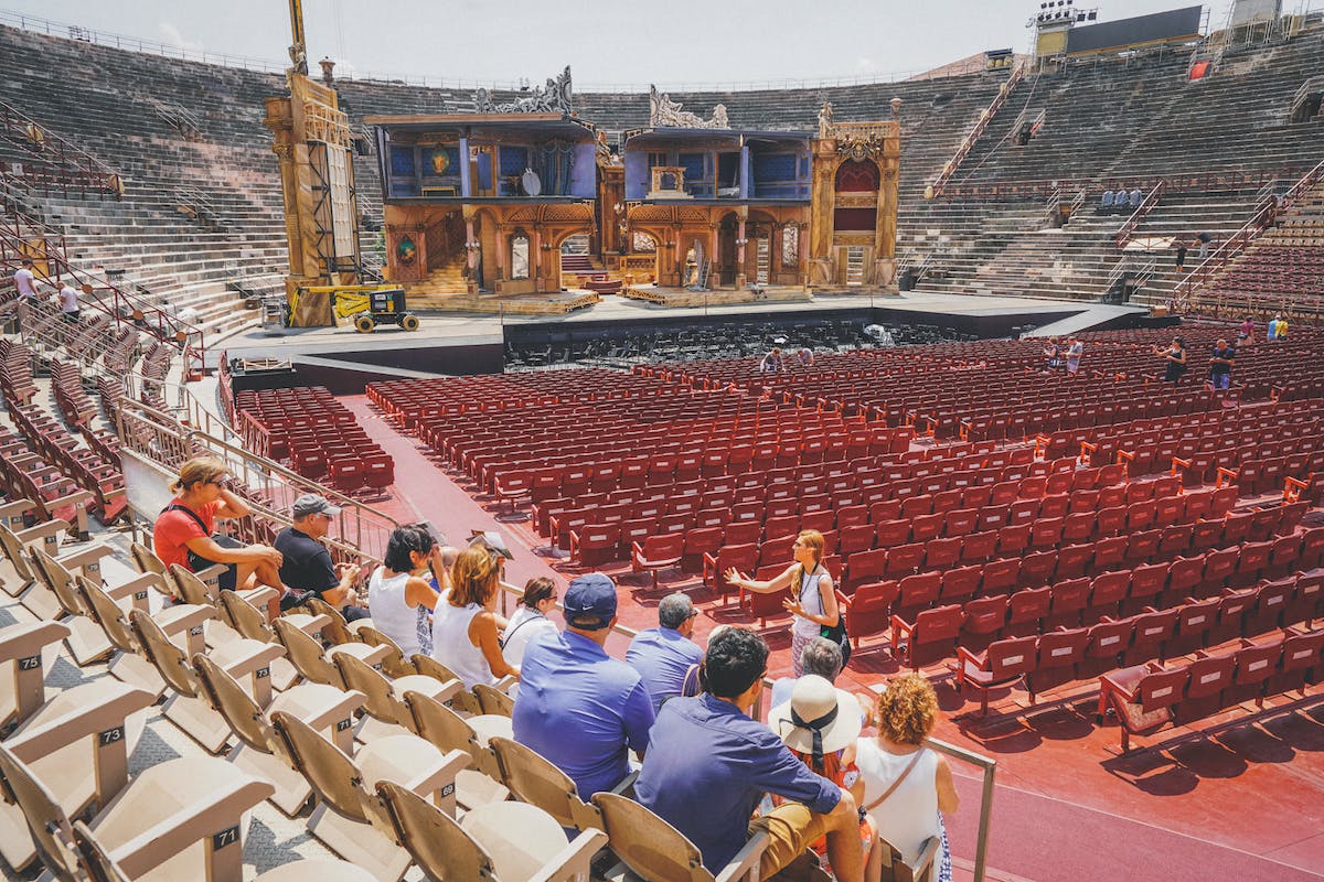 People seated in a large outdoor amphitheater facing an ornately decorated stage with empty red chairs in front.