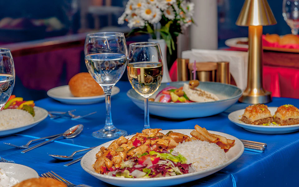 A table with plates of food, wine glasses filled with white wine, cutlery, and a vase of white flowers.