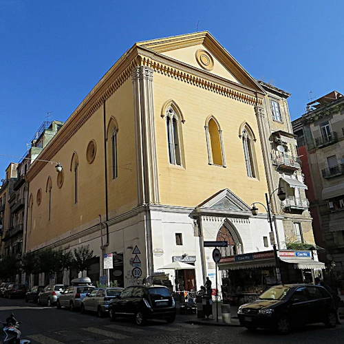 En stor gul byggnad med välvda fönster och en triangulär prydnad, placerad mellan andra byggnader längs en gata med parkerade bilar.