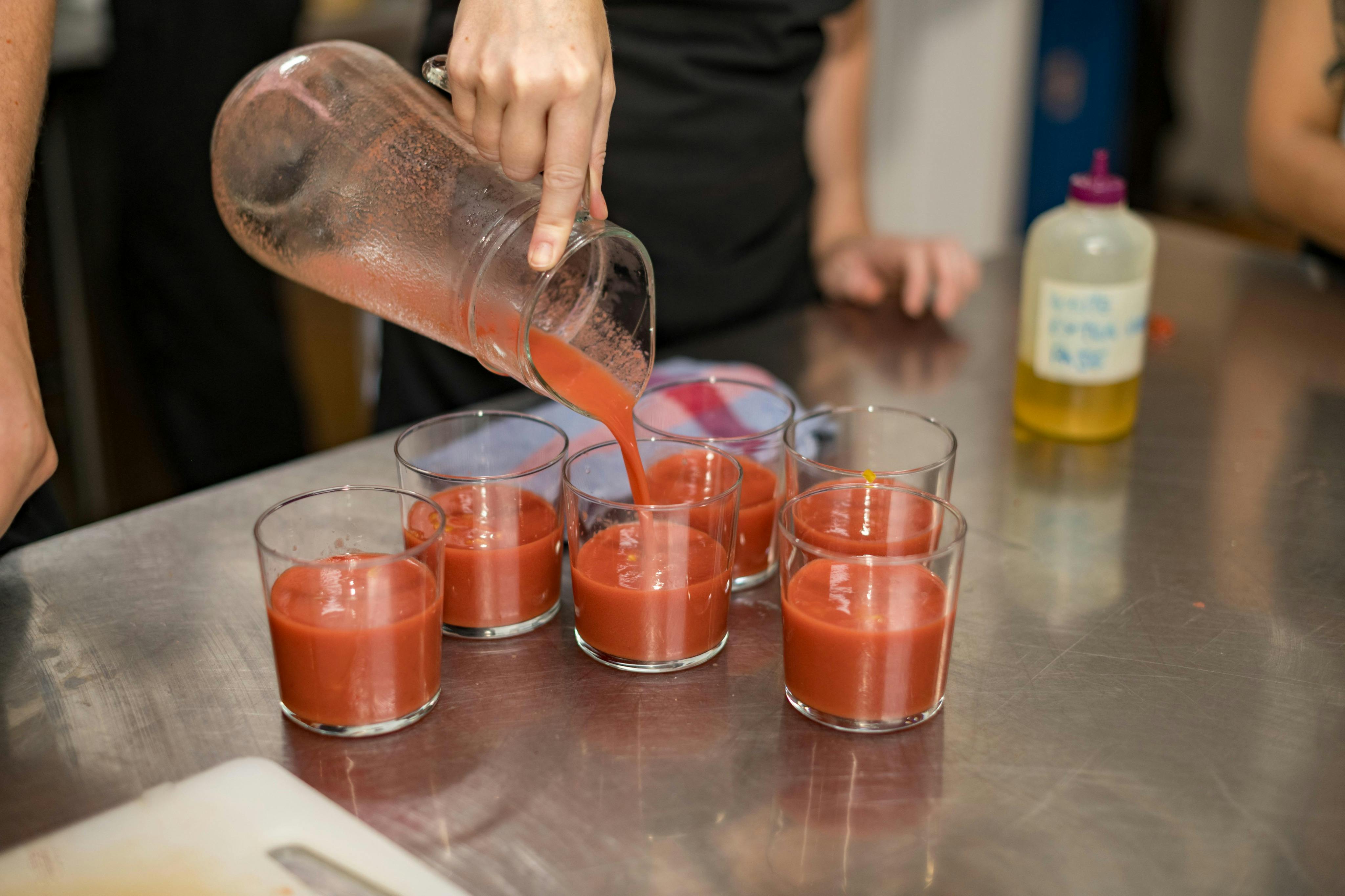 Ένα άτομο ρίχνει κόκκινο χυμό από μια κανάτα σε έξι ποτήρια σε έναν μεταλλικό πάγκο.