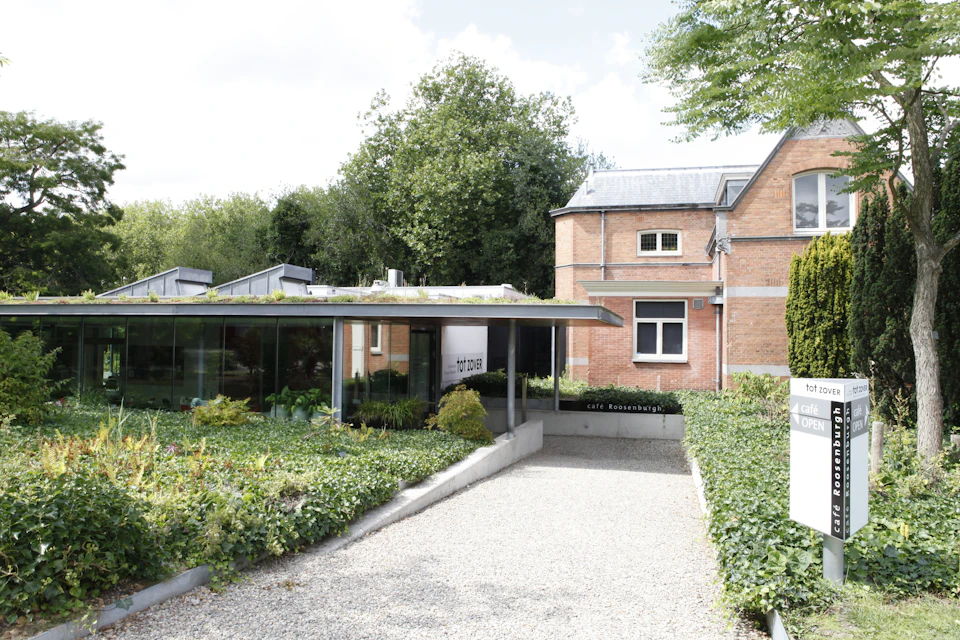 Brick building with modern glass extension, surrounded by greenery and trees. A pathway leads to an entrance labeled "Cafe Rozebroeken."