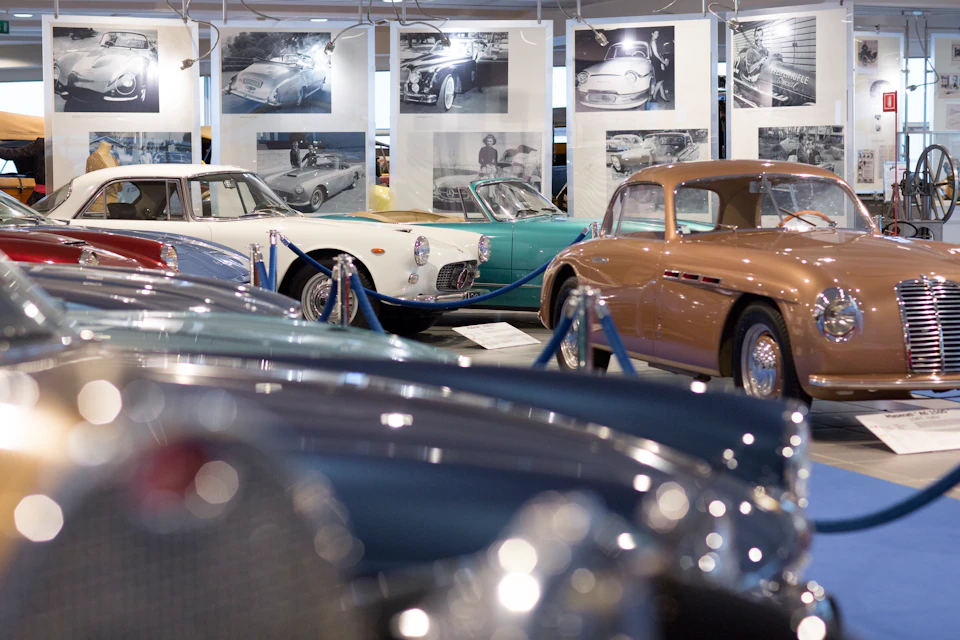 Classic car exhibit with various vintage cars displayed in a showroom, surrounded by photographs and roped-off sections.